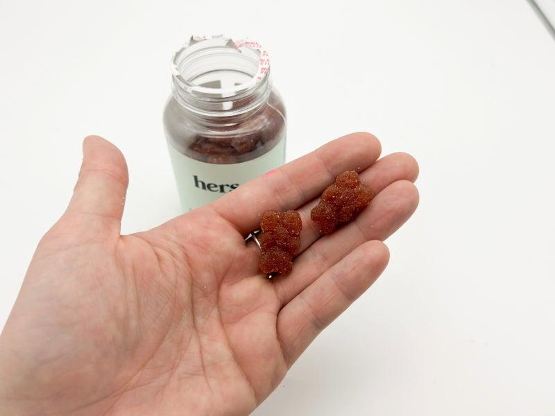 An open bottle of the Hers Multivitamin Gummy and two gummies sitting in a person's hand