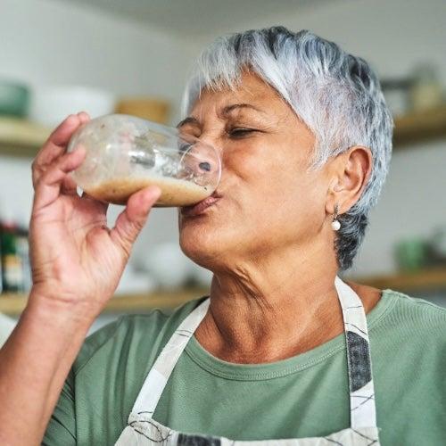 A woman with short grey hair drinks a protein shake from a glass