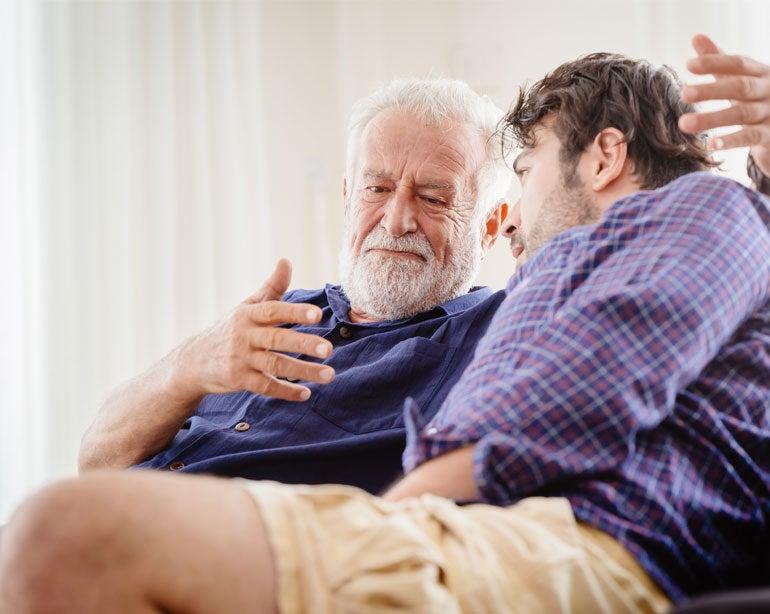 Older bearded man listens intently to a younger man as they sit close together, discussing in soft natural light.