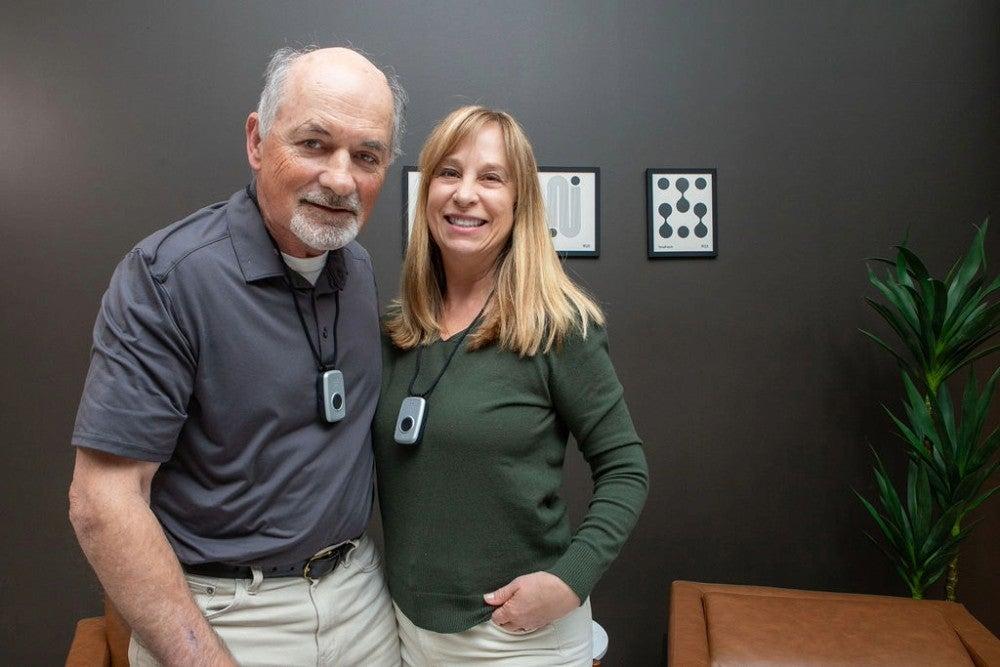 An older adult man and woman stand next to each other wearing medical alert necklaces