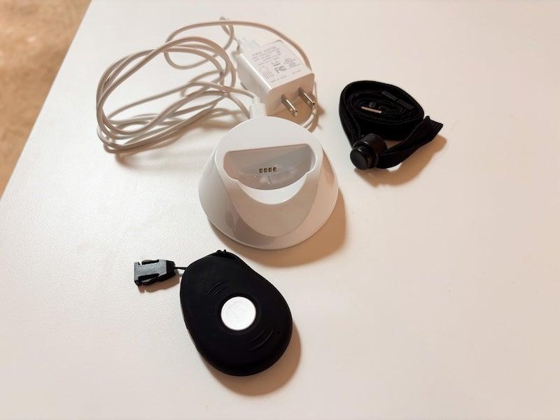 The LifeStation Sidekick, a black necklace the size and shape of a chicken nugget with a white dime-sized button in the center, sits beside its white charging cradle and a black lanyard that is rolled into a ball.