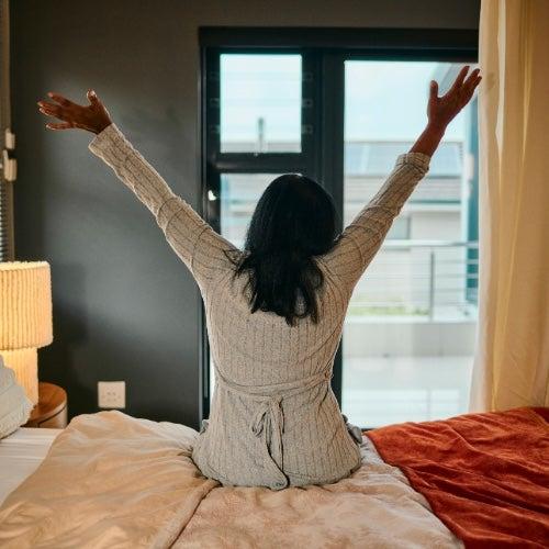 A woman sits on the edge of a bed and stretches her hands up