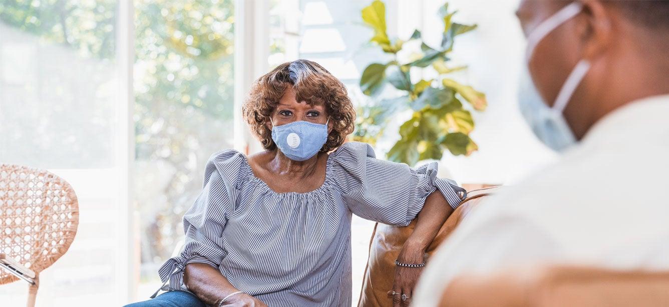 A senior Black older woman with blue mask is seated on a couch, talking to her mental health provider.
