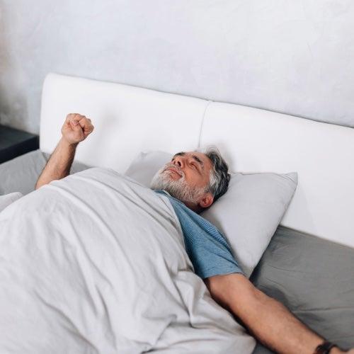 A man with grey hair and a blue shirt stretches in bed with his eyes closed
