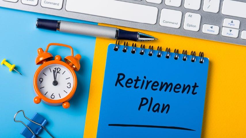 A blue spiral notebook with the words "Retirement Plan" on it sit atop a desk next to a clock, pen, push pin, large paper clip, and Mac keyboard.