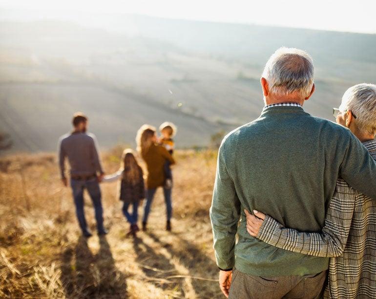 A senior couple hugs at the top of a hill, looking down at their family walking together.