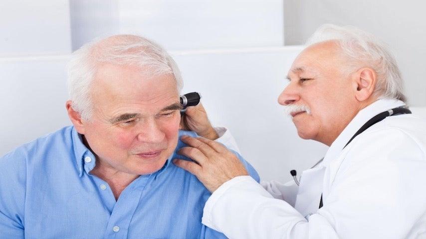 Male doctor wearing white lab coat examines the ear of an older adult man wearing a blue button-down shirt