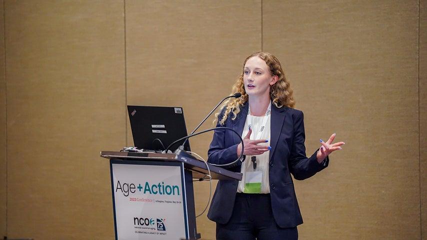 A person stands by a podium at the Age+Action conference, speaking to attendees in the audience.