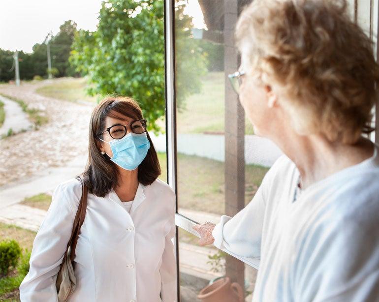 A female caregiver wearing a mask is standing at the door of a female senior's front door, paying her a visit/checking on her during the pandemic.