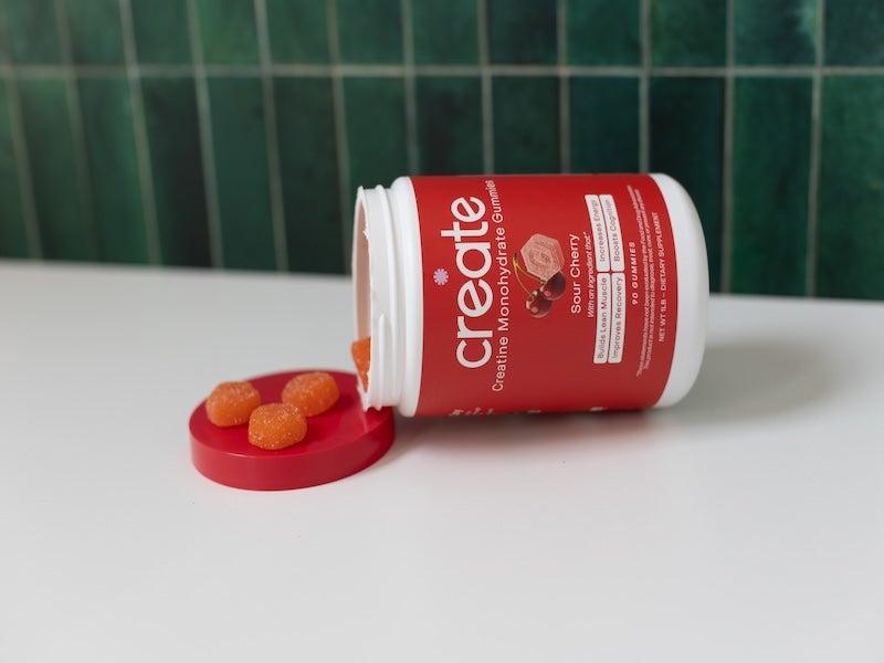 A red jar of Create Creatine Monohydrate Gummies lies on its side, spilling three gumdrops into its open lid