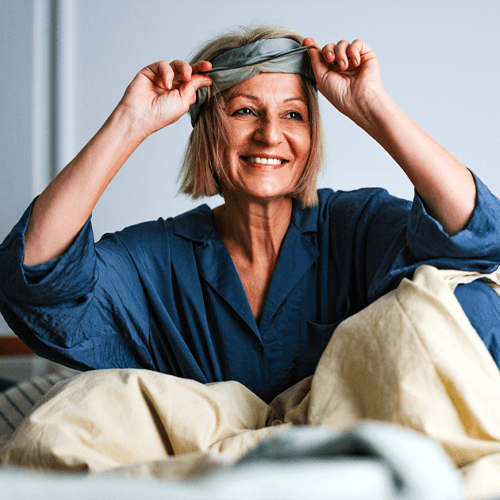 A woman sitting up in bed and pulling up a sleep mask while smiling