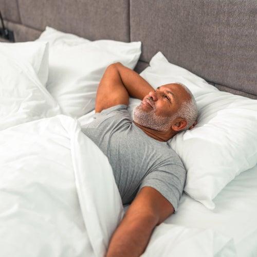 A man with short white hair and a white beard sleeps on a mattress with white sheets