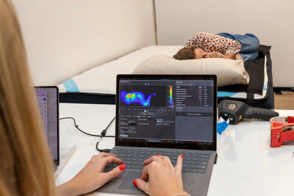 An older adult woman lying on her side on the DreamCloud Premier Memory Foam mattress, while another person shows a pressure map on a laptop