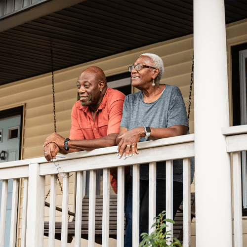 A couple leans against a porch railing and stares wistfully into the distance; she wears glasses and a grey shirt, he wears a persimmon shirt and is holding his glasses in his right hand.