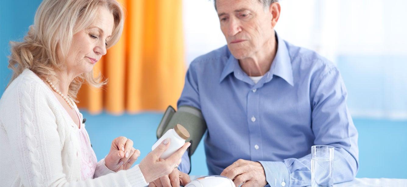 An older woman is supporting her senior husband while measuring blood pressure and looking at his medication.