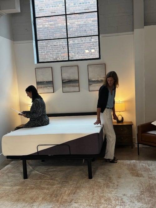 Two women testing a mattress by sitting on it and feeling its texture