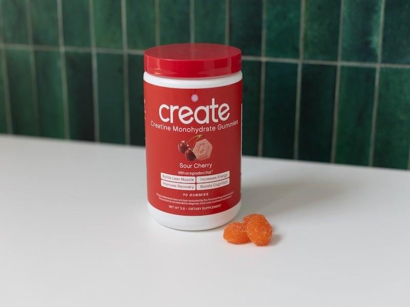 Red plastic jar of Create Creatine Monohydrate Gummies next to a pile of three burnt-orange gummies