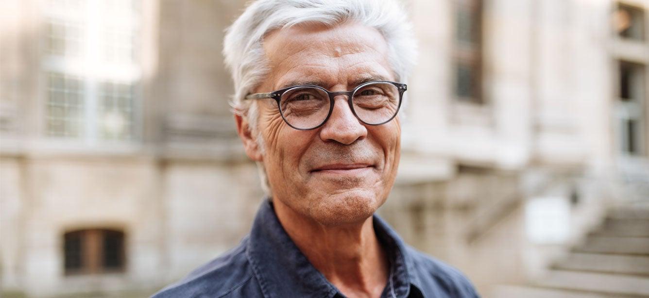Smiling older man with glasses and gray hair standing outdoors in front of a blurred building background.
