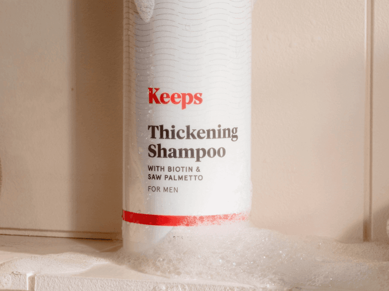 A bottle of Keeps Thickening Shampoo with soap suds around the bottom of the bottle