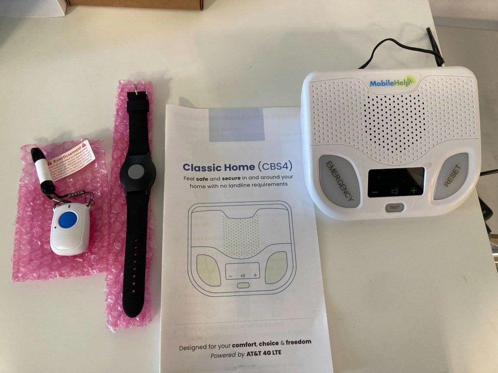The MobileHelp Classic Home system’s base station, user manual, help button in a wristband, and fall alert button sit on a table