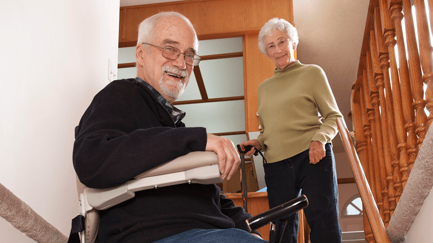 smiling older man riding stair lift as smiling older woman stands with cane at top of stairs