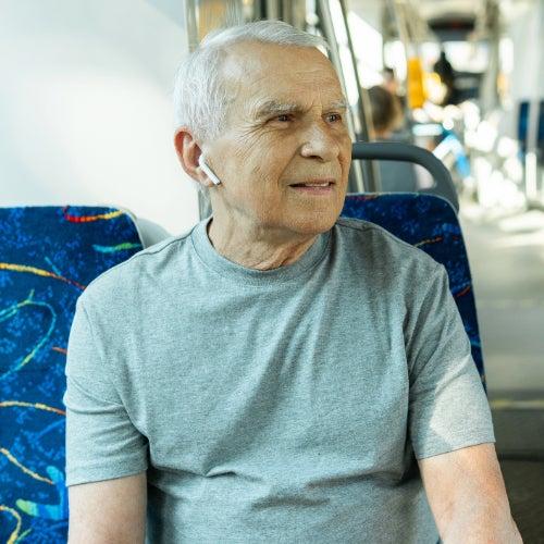A man with white hair sits on a bus with Apple AirPods earbuds in his ears