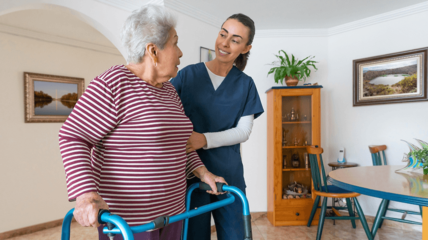 aid in medical scrubs holding arm of and smiling at older woman using walker