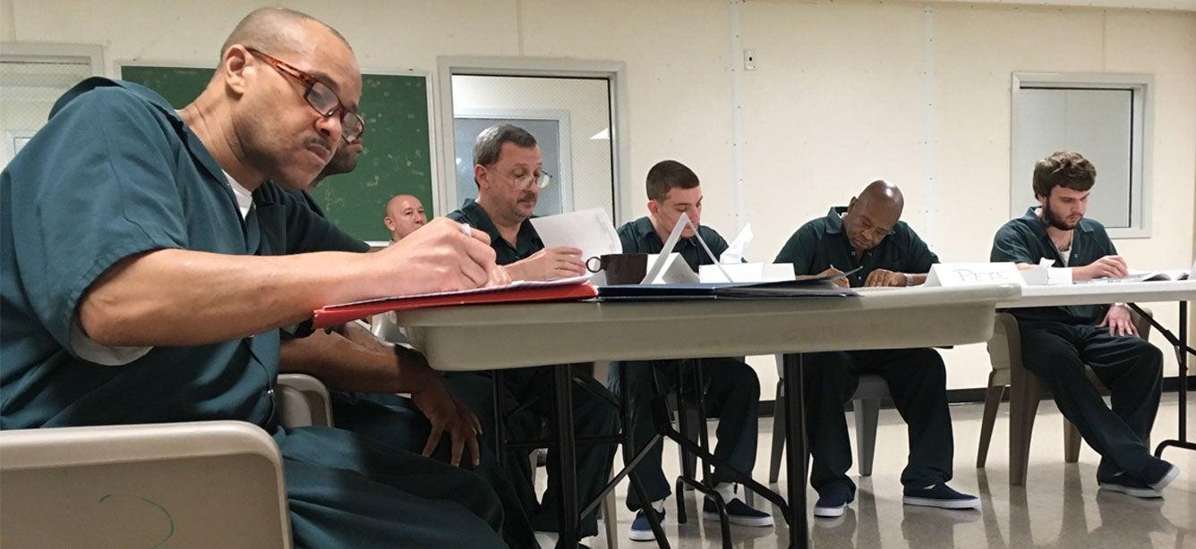 A group of prisoners of all ages are in a classroom setting filling out paperwork, studying together.