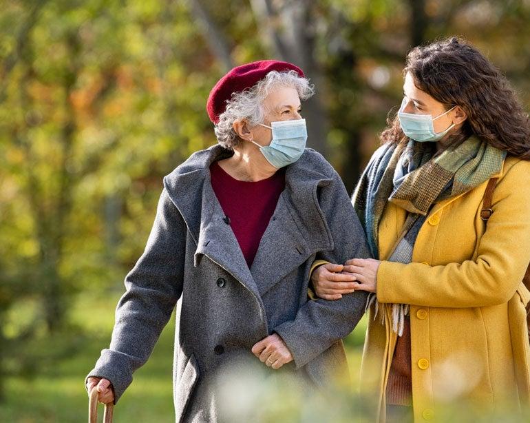 A senior Caucasian woman is walking with a younger adult outside while wearing a mask.