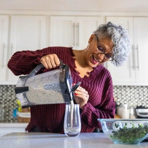 A woman with short, curly grey hair pours a smoothie into a glass in her kitchen