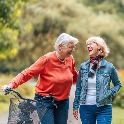 A woman with silver hair and an orange sweater walks next to a bicycle while laughing with her friend