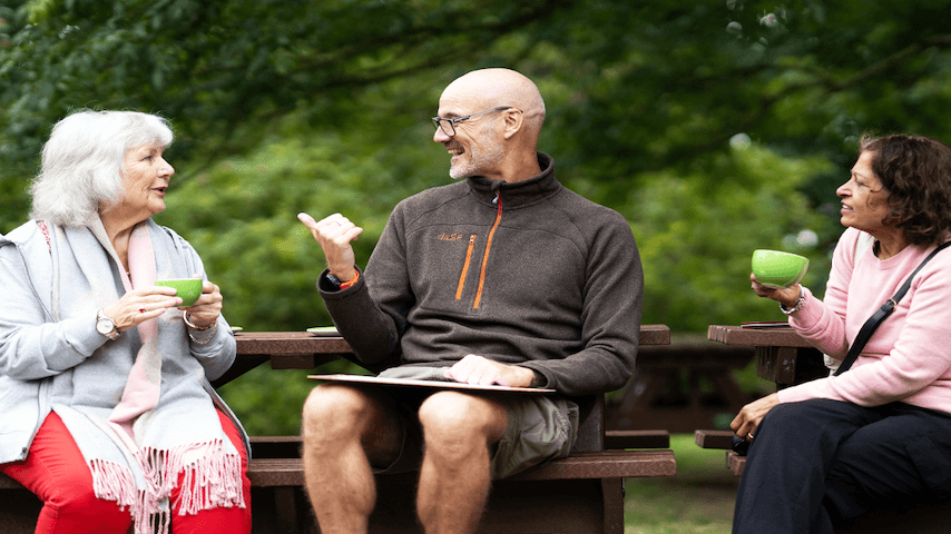 Three older adults chatting and drinking coffee at outdoor picnic table