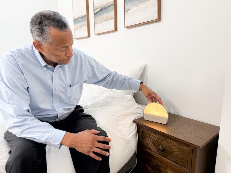 A man in a periwinkle button-down shirt sits on a bed to adjust the brightness of a Hatch Restore 3 sound machine alarm clock
