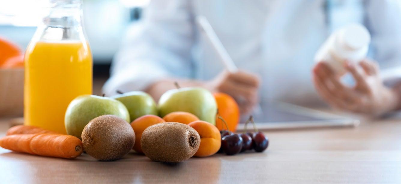 Fruits, juice, and carrots on a table, with someone holding a pill bottle and writing in the background.