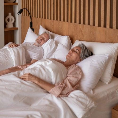 An older adult couple sleeps side by side in a bed with a wooden headboard