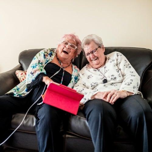 Two women sitting on a chair laughing while holding a tablet