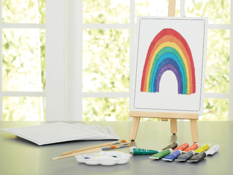 Canvas with a rainbow painted on it, sitting on a mini easel with paints and a palette on a table