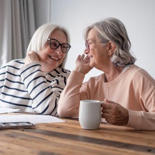 Two older women with silver hair sit at a table together and laugh