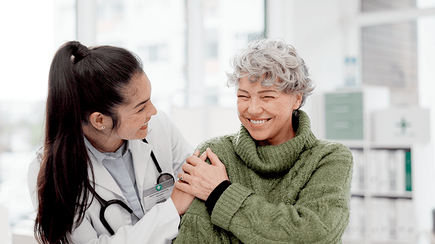 woman wearing lab coat and stethoscope laughing with older woman in green turtleneck sweater
