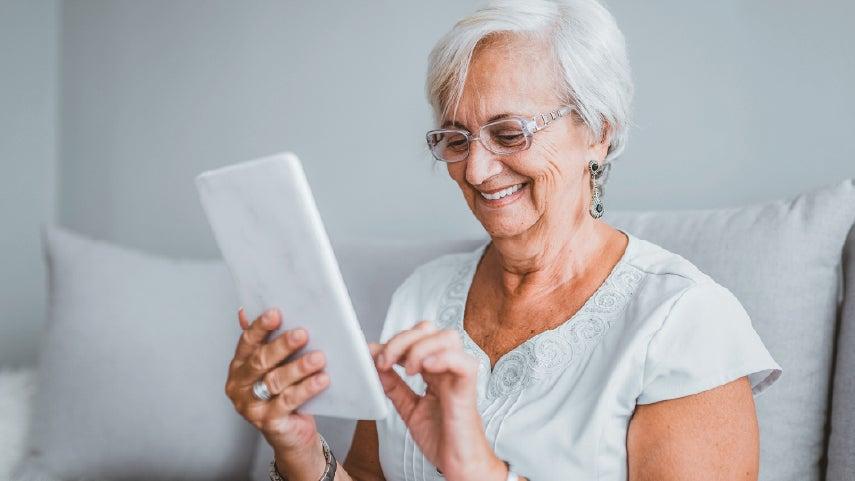 A senior caucasian woman with glasses is smiling while looking at her tablet.
