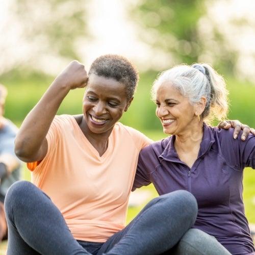Two older adult women laugh and flex their muscles as they participate in an outdoor workout group together