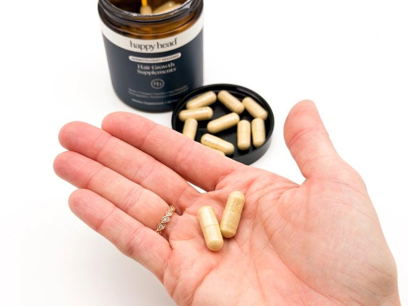 An open container of Happy Head Hair Growth Supplements, and two capsules in a person's hand