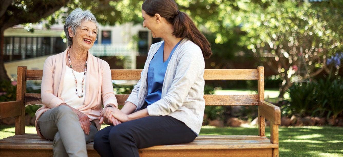 A senior woman and her caregiver are sitting together on a park bench, enjoying being outside.