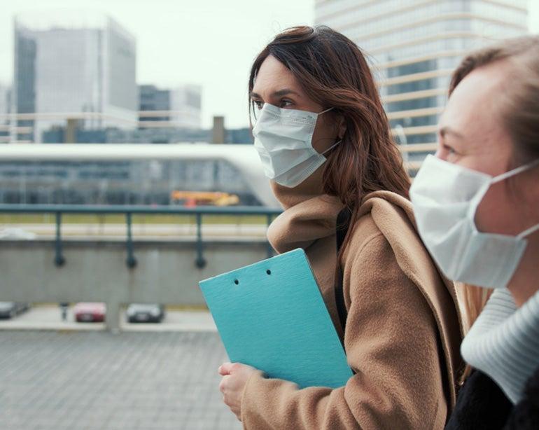 Two female professionals are walking together outside, one carrying a clipboard.