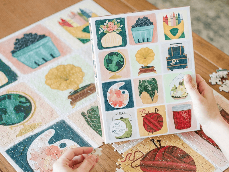 Hands hold a colorful puzzle box with twelve illustrated squares, showing items like a globe and crayons. The completed puzzle lies underneath