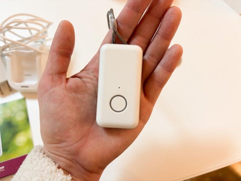 A white rectangular device is displayed in a person’s open left palm; the device is roughly as tall as a convenience store cigarette lighter, and two inches wide. Near the bottom of the device, there is a dime-sized white button.