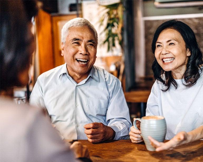 An Asian senior couple is sitting down having coffee is a coffee shop with their caregiver, loved one.