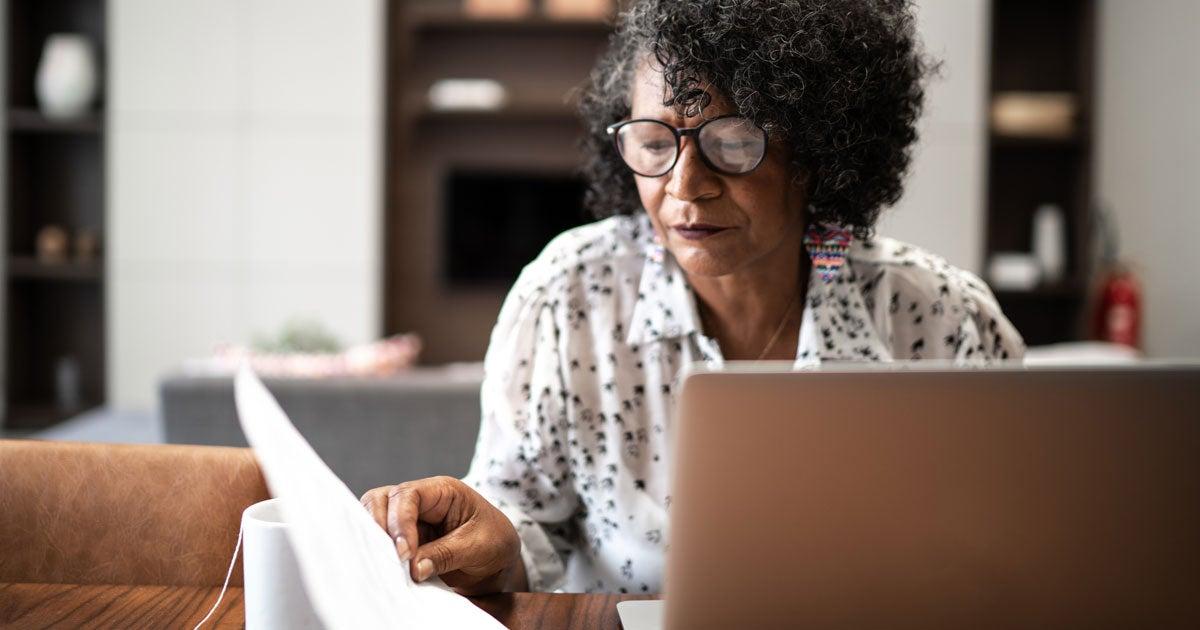 A Black senior woman is at her kitchen table, looking at her financials and working on her laptop.