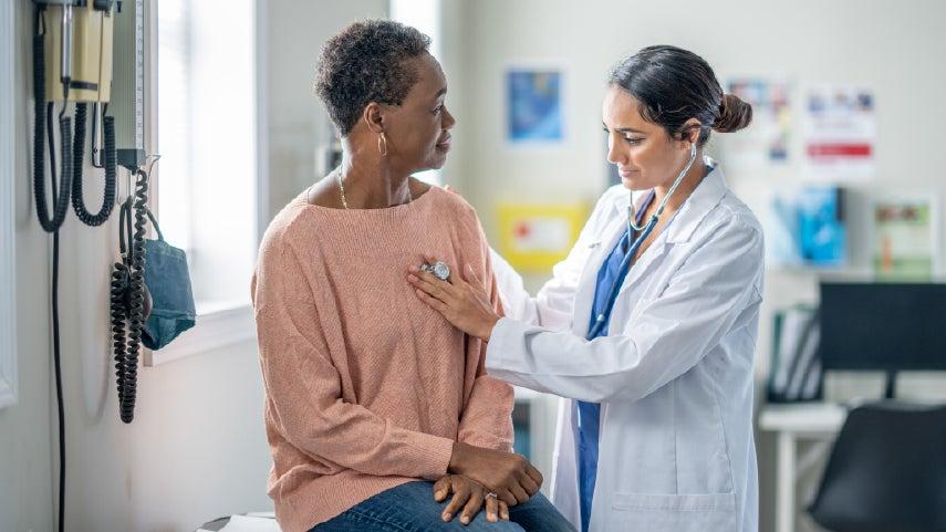 Woman sits while a medical professional checks her heart with a stethoscope.