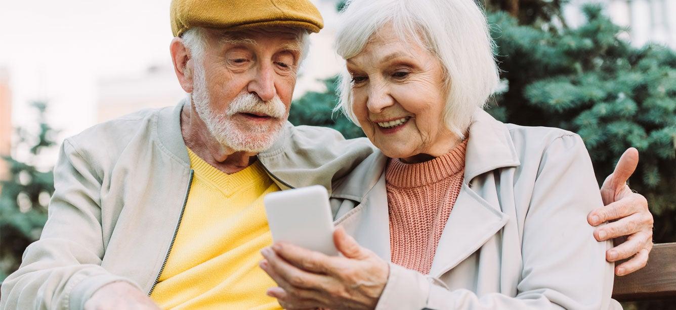 A senior Caucasian couple is sitting on a park bench video calling their child, smiling and enjoying conversation.